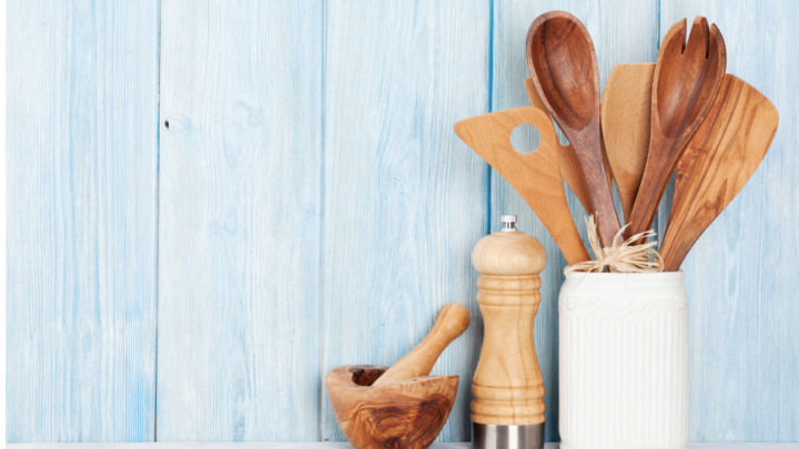 Close-up of essential kitchen gadgets made from wood: a set of cooking spoons and spatulas in a container, a grinder, and a mortar and pestle, all resting on a white countertop against a weathered blue wood panel background, perfect for a culinary blog or recipe site.