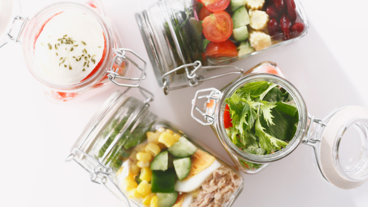 Healthy meal prep inspiration: an aerial view of four individual glass jars filled with different vibrant salads and one dessert. Each jar showcases distinct layers of vegetables, proteins, and fruits, set against a clean white surface with subtle shadows.
