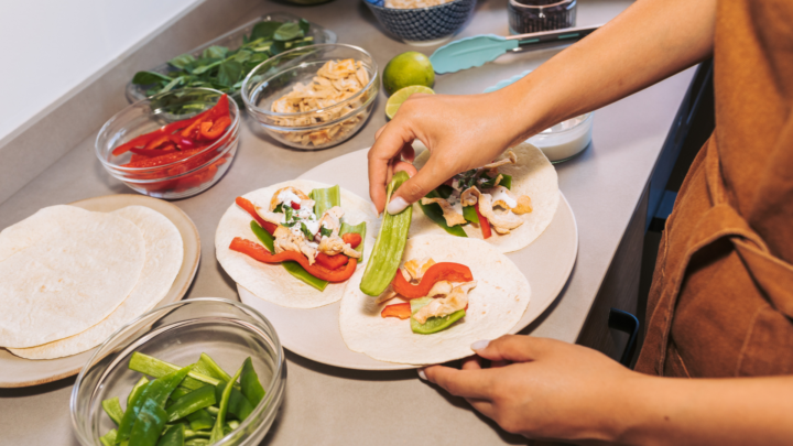 A person in a brown apron is assembling fresh, healthy wraps or tortillas with various colorful ingredients, including sliced chicken, bell peppers, and fresh greens. The scene captures the process of cooking or meal preparation in a bright kitchen.
