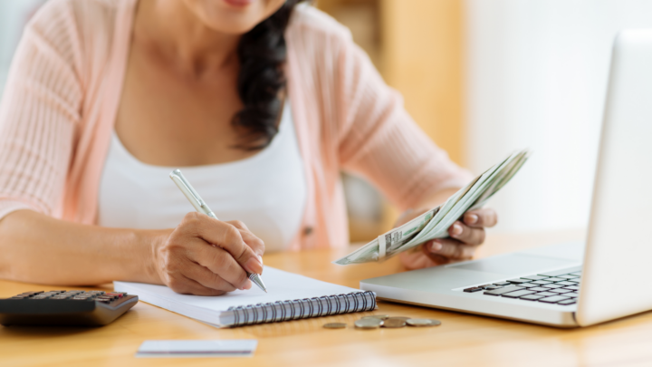 Close-up of a person managing personal finances, counting money, and making notes. The scene includes a calculator, laptop, and loose change, suggesting a focus on budgeting, savings, or business accounting.