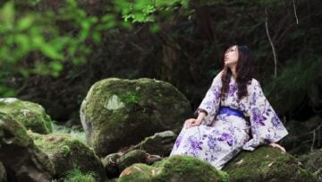 A woman in a purple and white floral kimono sitting on a mossy rock in a tranquil forest.