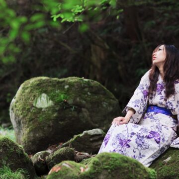 A woman in a purple and white floral kimono sitting on a mossy rock in a tranquil forest.
