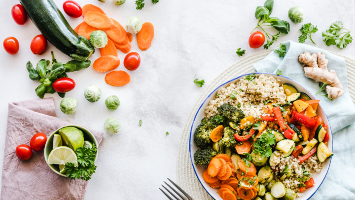 Fresh and wholesome ingredients for a balanced meal: a large bowl of cooked grains and mixed vegetables, with a composition of fresh zucchini, tomatoes, carrots, Brussels sprouts, and garnishes arranged artistically on a white table.